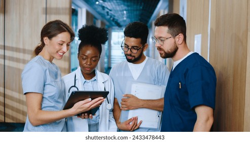 Multicultural group of youthful medical personnel communicating in modern hospital hallway during break. Nurses and doctors sharing experience and spend time using devices online. - Powered by Shutterstock