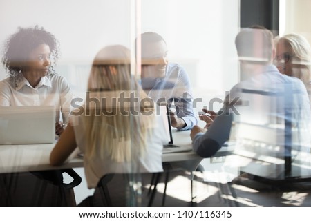 Multicultural international staff employees talking at team office company meeting sit at conference table behind glass door, diverse colleagues workers group collaborating at briefing in boardroom