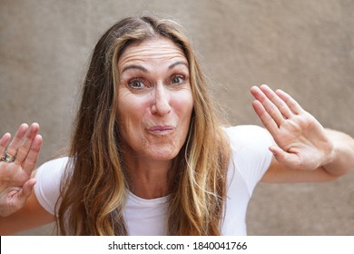 Multicultural Middle Aged Woman Making A Silly Face While Looking At Camera With Raised Eyebrows And Hands Up By Face For Closeup Portrait Outside Against Brown Stucco                               