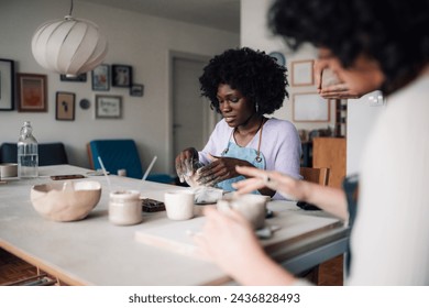 Multicultural pottery course students sitting in modern studio and learning clay work and arts and crafts together. Selective focus on african american woman doing pottery and making earthenware. - Powered by Shutterstock