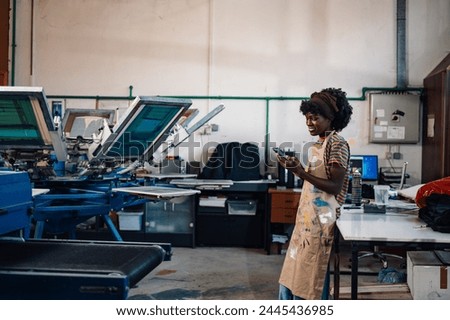 Multicultural printing shop worker at facility with cellphone in her hands and taking orders while standing near silkscreen machine. Smiling interracial small business owner using cellphone at factory