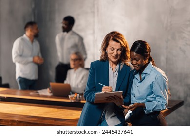 Multicultural professional businesswomen standing at office and writing down tasks and assignments in clipboard. Multiracial confident female managers writing down in clipboard and discussing project. - Powered by Shutterstock