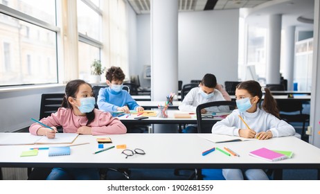 Multicultural schoolchildren wearing disposable medical masks sitting at table in classroom, keeping new normal social distance, looking at each other, studying at elementary school and writing - Powered by Shutterstock