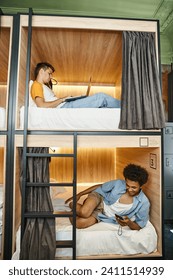 multicultural students using gadgets on double-decker beds in contemporary youth hostel, travelers