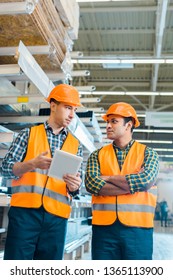 Multicultural Warehouse Workers In Safety Vasts And Helmets Talking While Using Digital Tablet