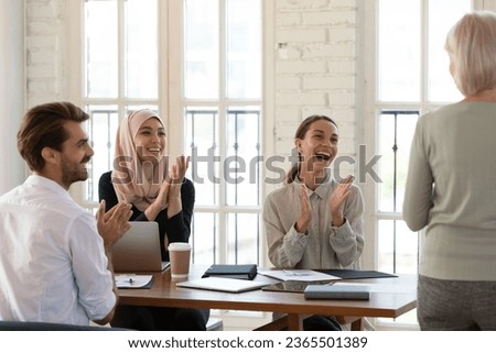 Multicultural young businesspeople celebrating successful negotiation accomplishment, grateful firm staff seated at boardroom table clap hands express appreciation to 60s middle-aged business trainer