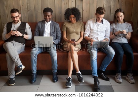 Multicultural young people using laptops and smartphones sitting in row, diverse african and caucasian millennials entertaining online obsessed with modern devices waiting in queue, gadget addiction