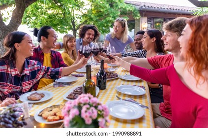 Multiethic Big Group Of People Enjoying Barbecue Dinner Together Outdoors Sitting On Garden Table Eating And Toasting Wine. Large Diverse Happy Friends Having A Summer Evening Picnic. Focus On Glasses