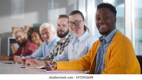 Multiethnic adult people sitting in row at desk and smiling at camera in classroom. Diverse people studying in adult education class or attending business seminar