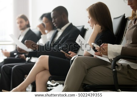 Multi-ethnic applicants sitting in queue preparing for interview, black and white vacancy candidates waiting on chairs holding resume using smartphones, human resources, hiring and job search concept
