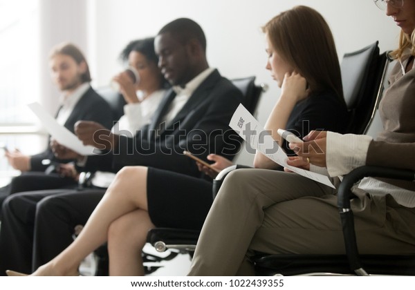 Multi-ethnic applicants sitting in queue\
preparing for interview, black and white vacancy candidates waiting\
on chairs holding resume using smartphones, human resources, hiring\
and job search\
concept