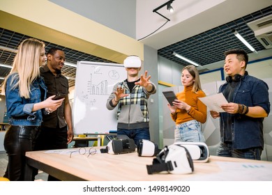 Multiethnic Business Team Presenting Virtual Reality Headset In Office Meeting For Young Handsome Man Chief. Developers Meeting With Virtual Reality Simulator Around Table In Creative Office