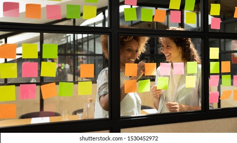 Multi-ethnic businesswomen friendly mates discuss business ideas and plans sharing thoughts writing on colourful sticky notes on transparent wall in office. Brainstorm and cooperation activity concept