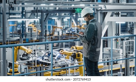 Multiethnic Car Factory Engineer In Work Uniform Using Tablet Computer. Automotive Industrial Manufacturing Facility Working On Vehicle Production With Robotic Arms. Automated Assembly Plant.