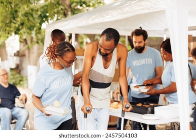 Multiethnic charity group handing out free food and nourishments to less fortunate and disabled. Portrait of black woman with blue t-shirt assisting the poor, needy african american man on crutches.