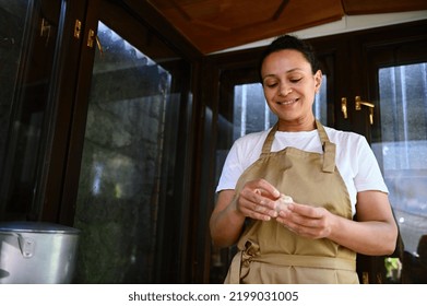 Multi-ethnic charming woman, a housewife cook wearing a beige chef's apron, hands-on, making homemade dumplings stuffed with mashed potatoes, according to traditional recipe, in the country kitchen - Powered by Shutterstock