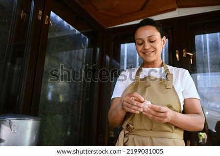 Multi-ethnic charming woman, a housewife cook wearing a beige chef's apron, hands-on, making homemade dumplings stuffed with mashed potatoes, according to traditional recipe, in the country kitchen