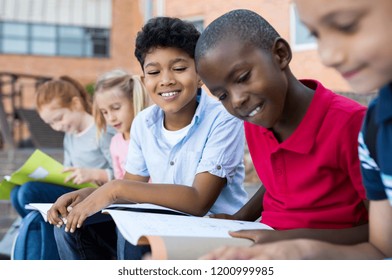 Multiethnic Children Sitting In A Row And Reading From Notebook All Together. School Kids Revising Notes For Exams Sitting On The Steps Outside The Elementary School. School Boy Studying For Classwork