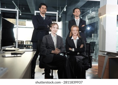 Multiethnic colleagues in dark suits pose together, the tan-skinned and bearded men standing while the two women sit smiling in a dim glass-walled workspace in a bright modern coworking.