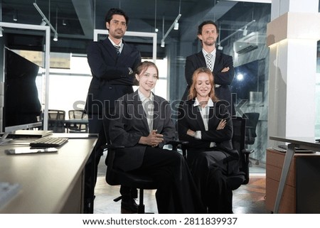 Multiethnic colleagues in dark suits pose together, the tan-skinned and bearded men standing while the two women sit smiling in a dim glass-walled workspace in a bright modern coworking.