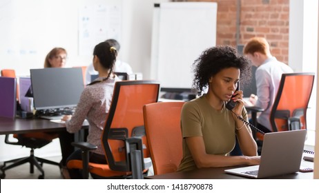 Multi-ethnic company employees sitting in modern shared office room sitting at desk use computer focus on black worker talking on landline phone typing message, busy workday in coworking space concept - Powered by Shutterstock