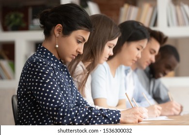 Multi-ethnic concentrated students sitting at desk holding pencils writing on papers doing task, diminishing perspective focus on indian girl study process or assessment - knowledge evaluation concept