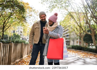 Multiethnic couple in love walking on the street in the city after buying christmas presents, using smartphone watching social media, having a good time together in winter season. Copy space. - Powered by Shutterstock