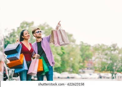 Multiethnic Couple With Shopping Bags, Man Pointing At Copy Space On Sky. Love, Casual Lifestyle, Or Shopaholic Concept