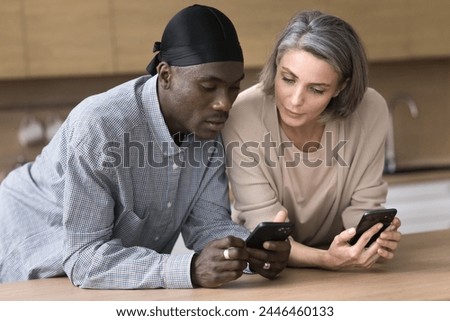 Multi-ethnic couple spend time in kitchen, stand at table with smartphones, exchange data between devices, share photo or contact, focused husband show new software application on cellphone to wife