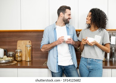 Multi-ethnic couple stands with a mugs in the kitchen and looks at each other with a love. An African woman and a caucasian guy enjoy morning coffee together at home - Powered by Shutterstock