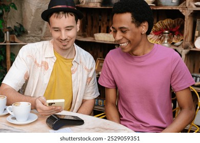 A multi-ethnic couple of two young men pay the bill in a coffee shop with a smartphone. They smile. The scene is casual and light-hearted. - Powered by Shutterstock