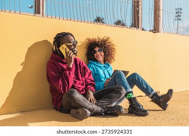 Multiethnic couple using mobile and tablet sitting against a yellow wall in urban setting outdoors