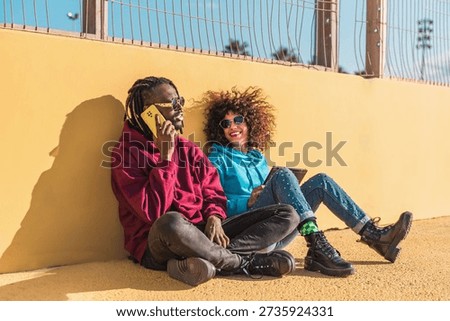 Multiethnic couple using mobile and tablet sitting against a yellow wall in urban setting outdoors