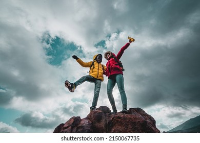 Multi-ethnic Couple Of Young People Hikers Excited On The Top Of The Mountain - View From Bottom Over A Cloudy Sky Background - Copy Space - Travel, Trekking And Lifestyle Concept