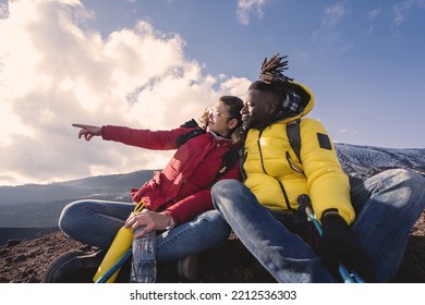 Multiethnic Couple Of Young People Relaxing On The Top Of The Mountain Sitting On The Floor And Pointing Fingers Looking Away - Wanderlust Hiker People And Diverse Couple Lifestyle Concept