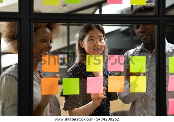 Multiethnic creative diverse young business team\
people three colleagues students group brainstorm on work plan talk\
write ideas tasks on post it sticky notes look at glass at scrum\
teamwork briefing