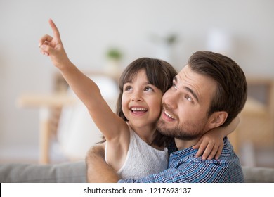 Multi-ethnic diverse happy family child and daddy sitting on couch together in living room at home. Preschool adorable positive daughter embracing with young father pointing with her hand upper left - Shutterstock ID 1217693137