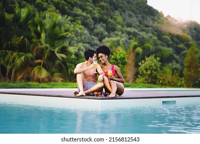 Multiethnic engaged couple sitting by the swimming pool drinking fruit mocktails in the summer surrounded by green hills - people relaxing on vacation lifestyle concept - Powered by Shutterstock