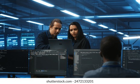 Multiethnic engineering team in server hub monitoring data center energy consumption across hardware using laptop. Multiracial colleagues inspecting supercomputers, ensuring malfunctions are fixed