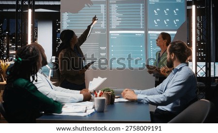 Multiethnic experts team evaluating data performance metrics on projection board, reviewing data insights with investors group in executive session after hours. Corporate event at night. Camera A.