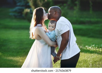 Multiethnic Family. Caucasian Mom And African American Dad Kiss And And Hold In Arms Small Child. Parents, Portrait Of Mom, Dad And Baby On Hands Over Green Grass Nature Background