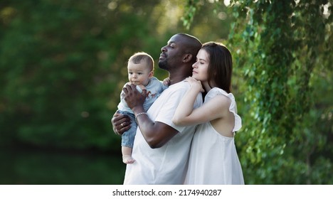 Multiethnic Family. Caucasian Mother And African American Father Hugging Child. Parents, Portrait Of Mom, Dad And Baby On Hands Over Green Nature Background