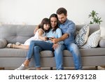 Multi-ethnic family sitting on sofa in living room at home. Mother father and little daughter using smartphone watching recreational program funny videos online having videocall talking with friends