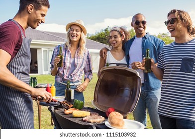 Multiethnic friends at barbecue party in the garden of house. Young man cooking meat on barbeque during summer holiday. Girls and guys having fun and drinking beers around the bbq. 
