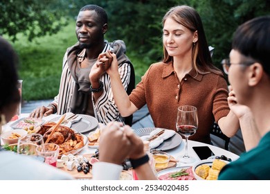 Multiethnic group of adult people holding hands at dinner table with eyes closed and saying grace during Thanksgiving celebration with friends outdoors - Powered by Shutterstock