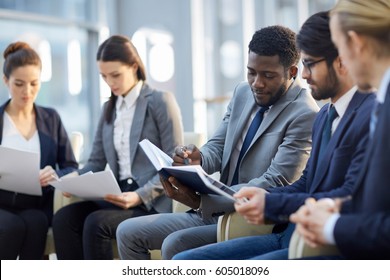 Multi-ethnic group of  business people sitting in row in modern glass hall, focus on African-American businessman reading handout materials and making notes in book - Shutterstock ID 605018096
