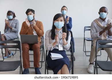 Multi-ethnic Group Of Business People Wearing Masks And Applauding With Social Distancing While Sitting On Chairs In Audience At Business Conference Or Seminar