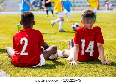 Multiethnic Group Of Children Playing Soccer Football Game. Two Young Multiracial Boys Wearing Red Soccer Jerseys Sitting on Grass at Soccer Field and Supporting Teammates