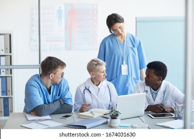 Multi-ethnic group of doctors communicating while sitting at desk with laptop in clinic office interior, copy space