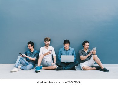 Multiethnic Group Of Four Men Using Smartphone, Laptop Computer, Digital Tablet Together With Copy Space On Blue Wall. Lifestyle With Information Technology Gadget Education Or Social Network Concept.